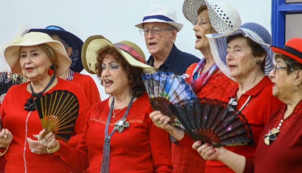 מקהלה - ששה נשים וגבר עם כובעים לראש ומניפה ביד , הנשים לבושות אדום הגבר בחולצה כחולה ושרים