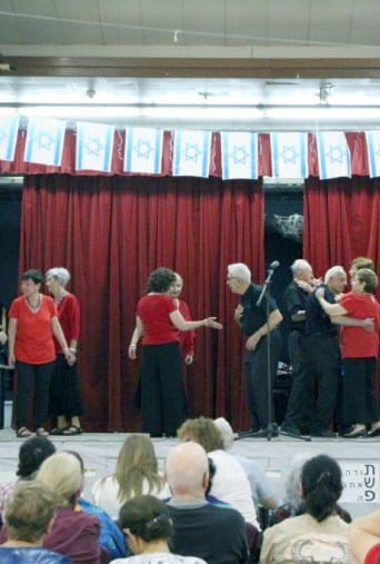 קבוצת אנשים כשהנשים לבושות בחולצות אדומות והגברים בשחור עומדים בקבוצות על הבמה וממול יושב קהל