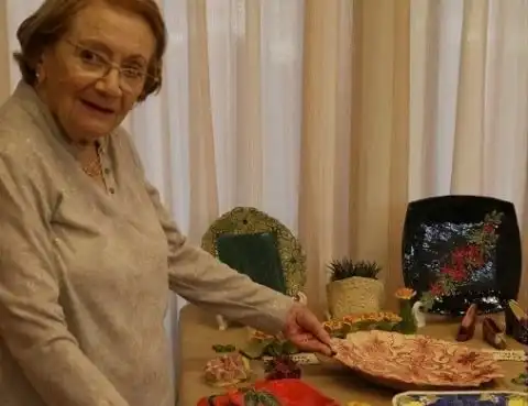 אישה עומדת ליד שולחן ומציגה חפצים :צלחות מקושטות , נעלים קטנות, עציך עם קקטוס ועוד